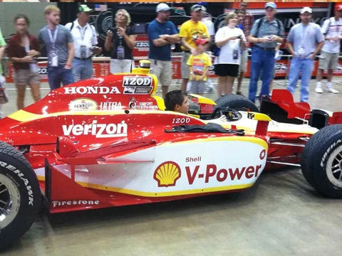 Red and yellow open-wheel race car beside spectators.
